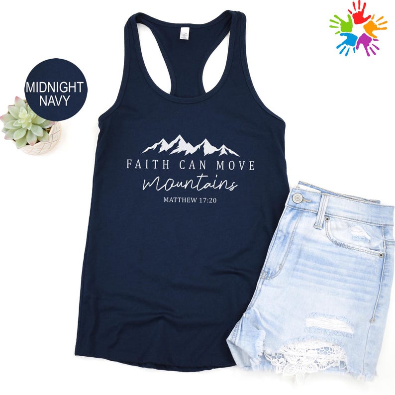 a navy blue tank top with a mountain range design and the text "Faith can move mountains" printed on it, along with the date "MATTHEW 17:20". Next to the tank top is a pair of denim shorts, and there is a small potted plant and a colorful handprint graphic in the background.