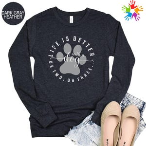 Może przedstawiać: Ciemnoszara melanżowa koszulka z długim rękawem z napisem "LIFE IS BETTER with a dog OR TWO, OR THREE." Projekt zawiera szary odcisk łapy. Koszulka jest połączona z dżinsowymi szortami i beżowymi balerinami.