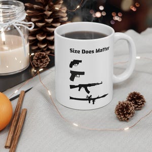 May include: White ceramic mug with black text and gun silhouettes. The mug reads "Size Does Matter" above a series of gun illustrations. The mug is filled with a dark liquid and sits on a light grey surface.