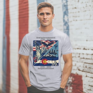 Può includere: T-shirt grigio chiaro con un design grafico che celebra il semi-quincentenario. Il design include la bandiera americana, la Statua della Libertà, un'aquila e la bandiera dello stato del Colorado. Il testo sulla maglietta recita "Semiquincentennial 1776-2026" e "1876-2026 Sesquicentennial."
