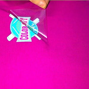 May include: A hand peels a clear transfer sheet from a vibrant magenta surface, revealing a colorful logo. The logo features the text "COLOR ART" in a pink and white design, set against a turquoise and white circular emblem.