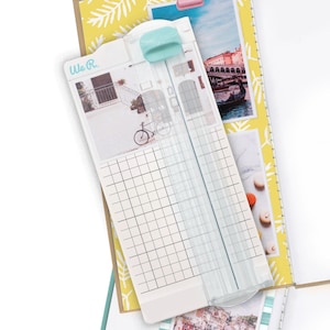 May include: A white paper trimmer with a mint green blade handle, resting on a scrapbook page. The trimmer has a grid for precise cuts and a photo of a bicycle. The scrapbook page has travel-themed photos.