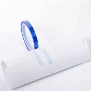May include: A roll of blue painter's tape is positioned above a white sheet of paper with two strips of tape applied. The tape is a vibrant blue color, and the paper is white. The image is well-lit, highlighting the tape's color and texture.