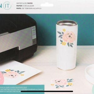 May include: A white tumbler with a floral design featuring pink and yellow flowers and green leaves. The image also shows a printer, water slide paper, and a printed A4 sheet with the same floral design.