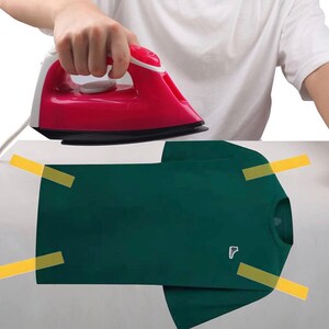 May include: A red and white iron being used on a folded dark green t-shirt. The t-shirt is secured with yellow tape. A small white logo is visible on the left chest area of the shirt. The person is wearing a white t-shirt.