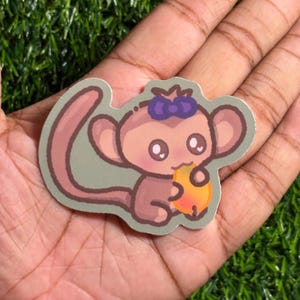 May include: A sticker featuring a cartoon monkey with a purple bow, holding a peach. The monkey is light brown with large eyes and a smiling expression. The sticker has a light green background and a white border.