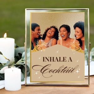 以下が含まれることがあります： 「Inhale a Cocktail」のテキストと、4人の女性が笑っている画像が入った額縁。フレームは金色で、背景は金色のアクセントが入った薄茶色です。2本の白いろうそくが前景にあります。