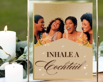 May include: A framed print with the text "Inhale a Cocktail" and an image of four laughing women. The frame is gold-colored, and the background is a light brown with gold accents. Two white candles are in the foreground.
