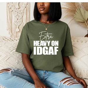 May include: Olive green t-shirt with the text "Extra Heavy On IDGAF" in white. The shirt has short sleeves and a crew neck. The model is wearing blue jeans with ripped knees.
