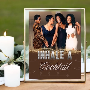Peut inclure: Impression encadrée avec quatre femmes en tenues élégantes, se tenant par la main et riant. L'impression porte les mots "INHALE A Cocktail" en lettres argentées et blanches sur fond marron, avec des icônes de voyage. Une bougie allumée et de la verdure sont au premier plan.