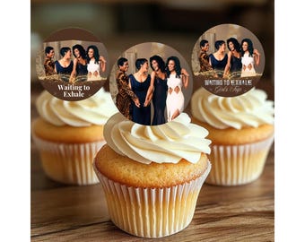May include: Three cupcakes with white frosting and edible toppers. The toppers feature images of women and the text "Waiting to Exhale" and "Girls Trip". The cupcakes sit on a wooden surface.