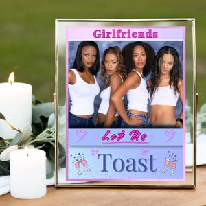 May include: A framed photo featuring four women in white tops and blue jeans. The photo has text that reads "Girlfriends" and "Let Us Toast". The frame is gold and sits on a wooden surface.