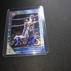 Puede incluir: Una tarjeta coleccionable de baloncesto NBA Hoops holográfica que muestra a Derrick Rose con el uniforme de los Detroit Pistons, en el aire durante una jugada. La tarjeta incluye un autógrafo en tinta azul al frente y está en una funda.