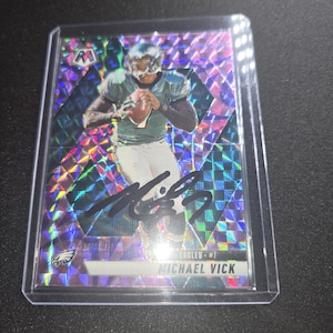 May include: A Michael Vick Philadelphia Eagles football trading card in a protective plastic sleeve. The card features a holographic purple and blue geometric pattern, a player in a team uniform holding a football, and a black ink autograph across the front.