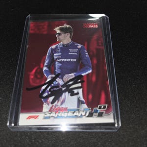 Puede incluir: Una tarjeta coleccionable rectangular con una fotografía de Logan Sargeant con el uniforme azul del equipo Williams Racing, usando gafas de sol y sosteniendo un casco. La tarjeta tiene un fondo rojo, un autógrafo en tinta negra y el texto Paddock Pass y F1.