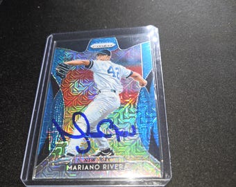 Puede incluir: Una tarjeta deportiva Prizm que muestra a Mariano Rivera con un uniforme de béisbol blanco con el número 42, capturado en un movimiento de lanzamiento. La tarjeta tiene un fondo geométrico azul y holográfico, un autógrafo en tinta azul y el texto Mariano Rivera.