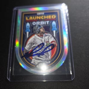 Può includere: Una figurina da baseball Topps Chrome intitolata Launched Into Orbit che raffigura un giocatore in divisa bianca con un autografo a inchiostro blu al centro. La carta ha un bordo olografico ed è inserita in una custodia protettiva trasparente.