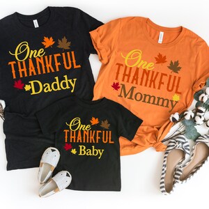 May include: Three t-shirts with the text "One Thankful Daddy", "One Thankful Mommy", and "One Thankful Baby" in orange and yellow script. The shirts are black and orange. Autumn leaves are printed above the text. A pair of shoes is next to the shirts.