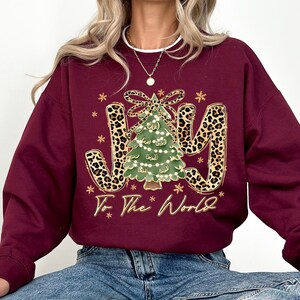 A woman wearing a burgundy sweatshirt with a Christmas tree design and the text "Joy To The World" printed on it.