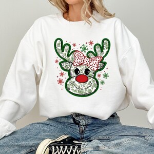 Könnte beinhalten: Weißes Sweatshirt mit einem Rentiergesicht-Design. Das Rentier hat grüne Geweihe, eine rote Nase und eine rot-weiß gepunktete Schleife. Das Gesicht des Rentiers ist mit einem floralen Muster verziert und von roten und grünen Schneeflocken umgeben.