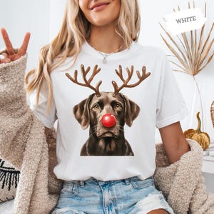 A woman wearing a white t-shirt with a graphic of a dog wearing antlers and a red nose, smiling and making a peace sign.