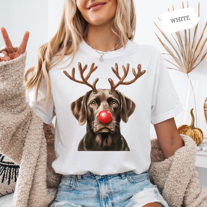 A woman wearing a white t-shirt with a graphic of a dog wearing antlers and a red nose, smiling and making a peace sign.