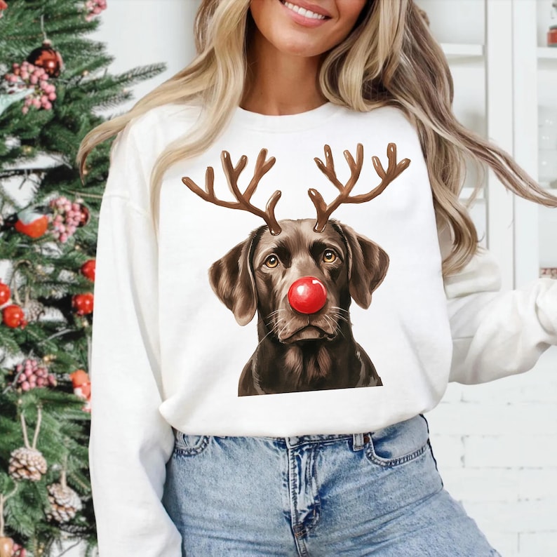 A woman wearing a white sweater with a graphic of a dog wearing antlers and a red nose, standing in front of a Christmas tree.