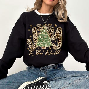 A woman wearing a black sweatshirt with a Christmas tree design and the text "To the World" printed on it.