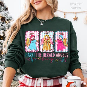 May include: Forest green sweatshirt featuring a graphic of three angels with halos and wings, each in a different colored dress. The text "Hark! The Herald Angels Sing" is below the angels, with musical notes. A festive holiday design.