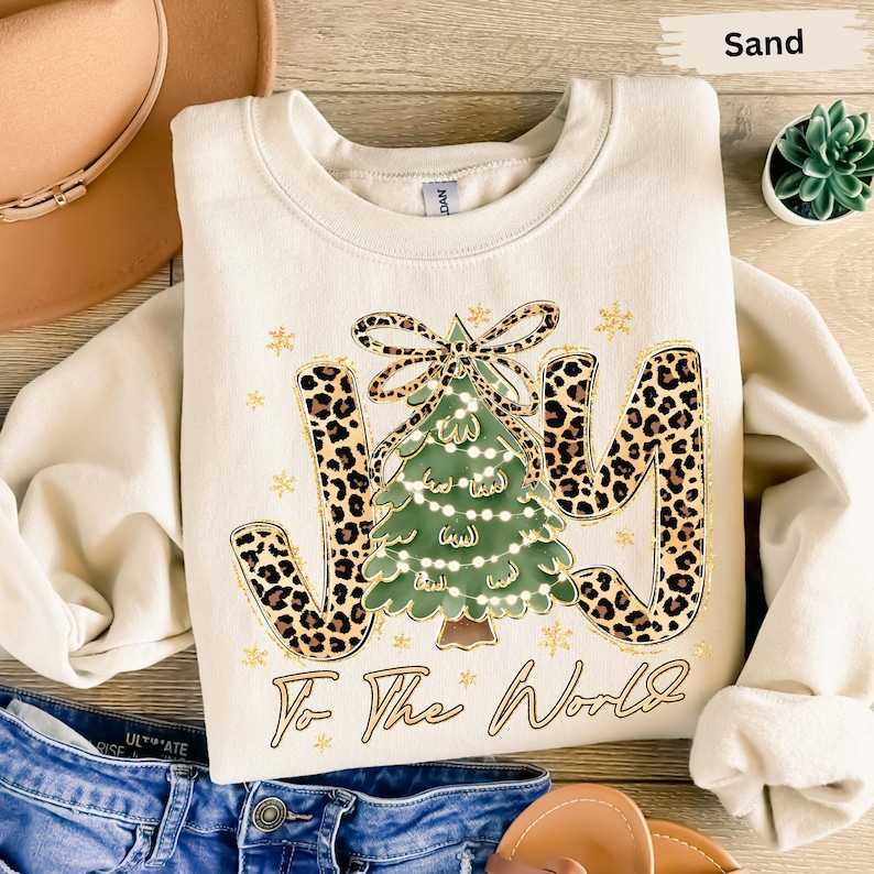 A white sweatshirt with a leopard print design and the text "Joy To The World" printed on it, along with a Christmas tree and snowflakes. The sweatshirt is placed on a wooden surface, and there are some other items such as a hat, a small plant, and a pair of jeans visible in the image.
