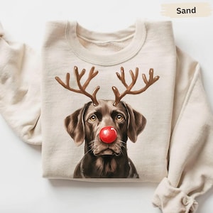 May include: Sand-colored sweatshirt featuring a brown dog with reindeer antlers and a red nose. The dog's face is the focal point, with detailed fur and expressive eyes. The antlers are brown, and the nose is a bright red. The sweatshirt is a neutral color.