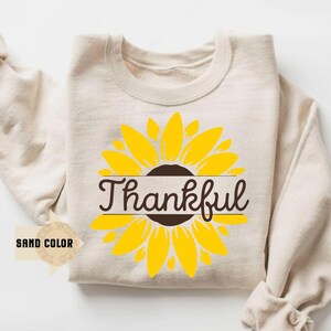 May include: A sand-colored sweatshirt featuring a large yellow sunflower design with a brown center. The word "Thankful" is written in a brown script across the center of the sunflower. The sweatshirt has long sleeves.