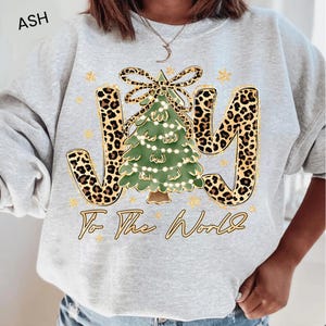 A person wearing a grey sweatshirt with a Christmas tree design and the text "Joy To The World" printed on it.