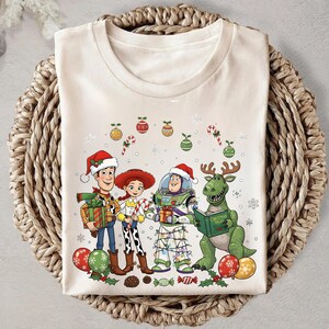 A white t-shirt with a Christmas-themed graphic design featuring characters from the Toy Story franchise, including Woody, Buzz Lightyear, and a dinosaur.