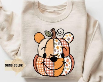 May include: A sand-coloured sweatshirt with a smiling pumpkin-bear design. The design features patchwork patterns in orange, cream, and floral prints. The bear-pumpkin has a stem and leaf detail at the top.