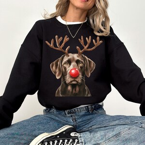 A woman wearing a black sweatshirt with a graphic of a dog wearing antlers and a red nose, sitting on a white background.