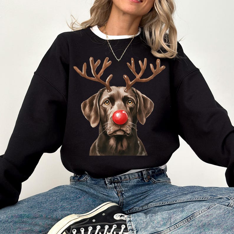 A woman wearing a black sweatshirt with a graphic of a dog wearing antlers and a red nose, sitting on a white background.