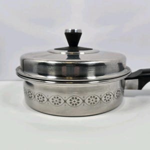 May include: Stainless steel saucepan with a lid and black handle. The pot has a decorative floral pattern around the base. The lid has a black handle. The pot is designed for cooking.