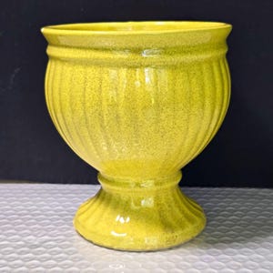 May include: A bright yellow ceramic planter with a speckled finish and vertical ribbed detailing. The vessel features a wide, flared rim, a rounded body, and a pedestal base with a matching ribbed design, creating a classic urn-like silhouette.