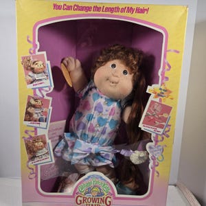 May include: Cabbage Patch Kids doll with growing hair in its original box. The doll has brown curly hair, freckles, and is wearing a blue, purple, and white heart-patterned dress. The box has text that reads "You Can Change the Length of My Hair!"