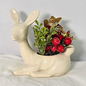 May include: A cream-colored ceramic deer figurine with a crackle glaze finish, featuring a hollow back filled with artificial green foliage, bright red berries, and a small decorative angel ornament wearing a red dress.