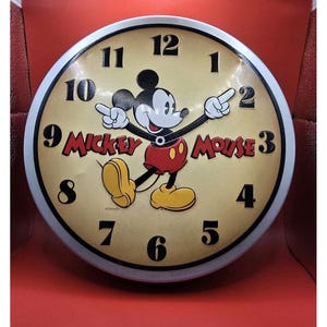 May include: Round wall clock featuring Mickey Mouse. The clock face is tan with black numerals and a white border. Mickey Mouse is in the center, with his arms outstretched, pointing at the numbers. The words "Mickey Mouse" are printed in red.