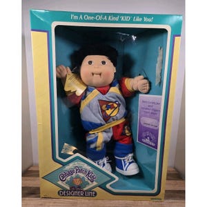 May include: A Cabbage Patch Kids Designer Line doll in its original box. The doll has black curly hair, a red and blue outfit, and white trainers. The box has text that reads "I'm A One-Of-A Kind 'KID' Like You!" and "Birth Certificate and Adoption Papers Application."