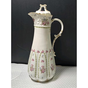 May include: A tall white porcelain pitcher featuring a delicate floral pattern of pink roses and blue flowers with green vine accents. The pitcher has a fluted body, a curved handle, and a matching lid topped with a decorative bow-shaped finial.