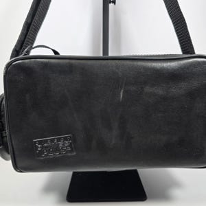 May include: Black leather shoulder bag with a black adjustable strap. The bag has a rectangular shape with a slightly textured surface. The words "POWER PLUS" are embossed on the lower front of the bag.