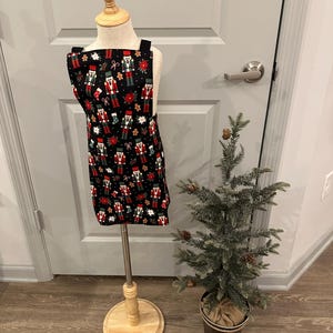Könnte beinhalten: Eine schwarze Schürze mit einem festlichen Weihnachtsmuster mit Nussknackern, Weihnachtssternen und Lebkuchenmännern. Die Schürze wird auf einer Holzpuppe präsentiert. Ein kleiner Weihnachtsbaum ist im Hintergrund.