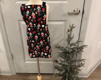 May include: A black apron with a festive Christmas-themed pattern featuring nutcrackers, poinsettias, and gingerbread men. The apron is displayed on a wooden mannequin. A small Christmas tree is in the background.
