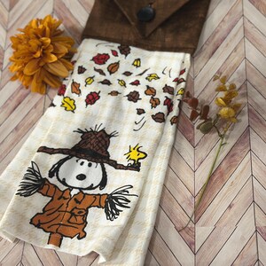 Könnte beinhalten: Ein dekoratives Küchentuch mit Snoopy als Vogelscheuche verkleidet mit Woodstock. Das Handtuch hat eine braune Oberseite mit Knopfverschluss und einen cremefarbenen Körper mit fallenden Blättern. Eine orangefarbene Blume und ein Zweig mit Blättern sind ebenfalls vorhanden.
