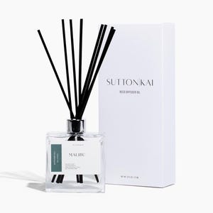 Könnte beinhalten: Ein klarer Glas-Schilf-Diffusor mit schwarzen Schilfrohren und einem silbernen Kragen. Der Diffusor ist mit "MALIBU" mit einem tealfarbenen Akzent beschriftet. Eine weiße Box mit dem Text "SUTTONKAI REED DIFFUSER OIL" steht daneben. Ein Artikel für Raumduft.