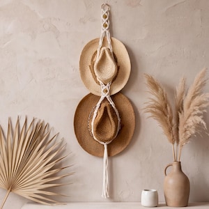 May include: A vertical macrame wall hanger made of off-white cord, featuring two circular loops designed to hold hats. The hanger is suspended from a wooden ring at the top and terminates in a long, knotted fringe at the bottom.